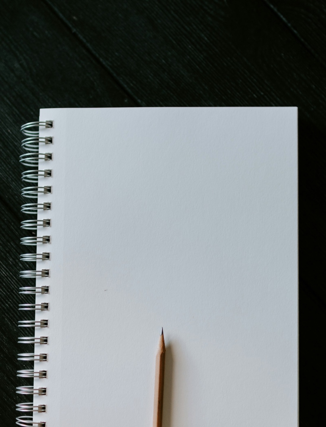 A blank spiral notebook and a sharpened pencil on a dark wooden table